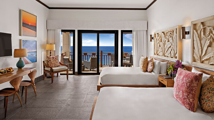 Duas camas queen size, mesa lateral com flores, cadeira, mesa e TV com vista para o mar da varanda