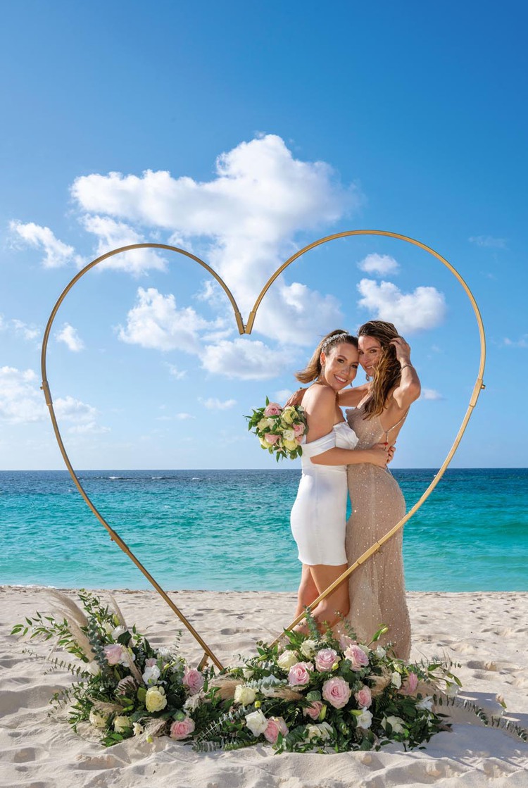Two women standing in front of ocean with wooden heart and flowers framing them